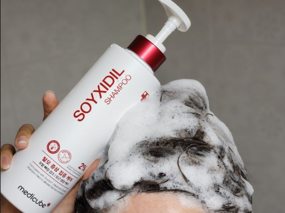 Person washing hair with SOYXIDIL shampoo, holding the bottle with a gray background